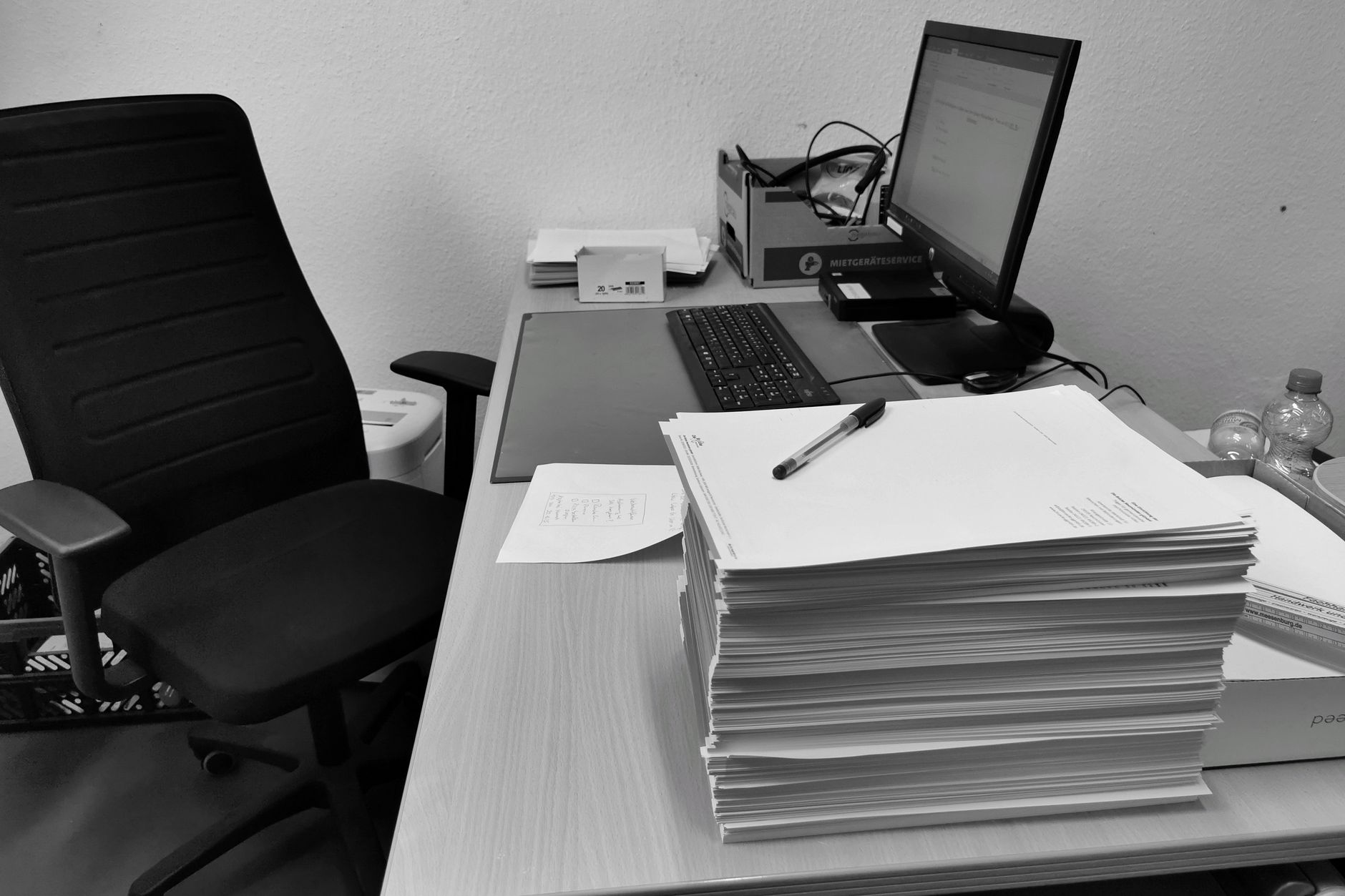 Black and white image of a cluttered office desk with a computer, chair, and paperwork.